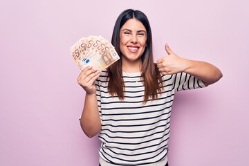 Young beautiful brunette woman holding euros banknotes over isolated pink background smiling happy and positive, thumb up doing excellent and approval sign