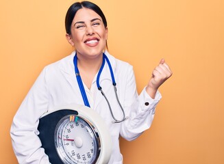 Young beautiful doctor woman wearing stethoscope and coat holding weighting machine screaming proud, celebrating victory and success very excited with raised arm