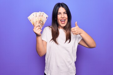 Young beautiful brunette woman holding euros banknotes over isolated purple background smiling happy and positive, thumb up doing excellent and approval sign