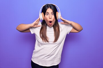 Young beautiful brunette woman listening to music using headphones over purple background scared and amazed with open mouth for surprise, disbelief face