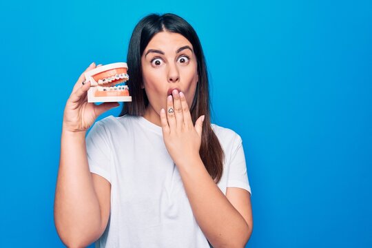 Young beautiful brunette woman asking for dental care holding denture with backets covering mouth with hand, shocked and afraid for mistake. Surprised expression