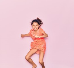 Adorable hispanic child girl on vacation wearing summer dress smiling happy. Jumping with smile on face over isolated pink background