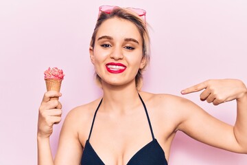 Young beautiful blonde woman wearing bikini holding ice cream pointing finger to one self smiling...