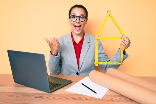 Young brunette architect woman at the office holding build project pointing thumb up to the side smiling happy with open mouth