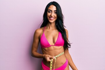 Naklejka premium Beautiful hispanic woman wearing bikini and measuring waist looking positive and happy standing and smiling with a confident smile showing teeth