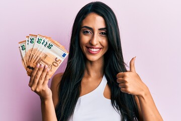 Beautiful hispanic woman holding 50 euro banknotes smiling happy and positive, thumb up doing excellent and approval sign