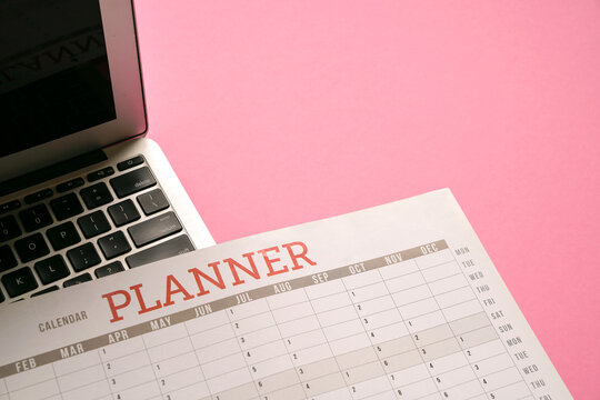 Calendar planner and computer laptop on top of pink table top.