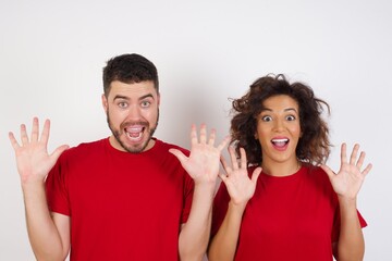 Optimistic Young beautiful couple wearing red t-shirt on white background raises palms from joy, happy to receive awesome present from someone, shouts loudly, Excited couple screaming.