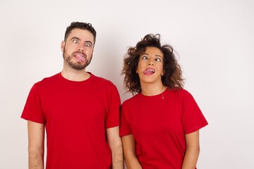 Young beautiful couple wearing red t-shirt on white background showing grimace face crossing her eyes and showing tongue . Being funny and crazy