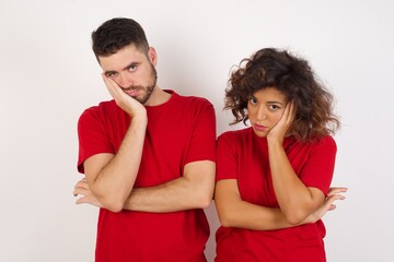 Very bored Young beautiful couple wearing red t-shirt on white background holding hand on cheek while support it with another crossed hand, looking tired and sick,