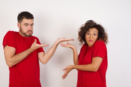 Young beautiful couple wearing red t-shirt on white background pointing aside with both hands showing something strange and saying: I don't know what is this. Advertisement concept.