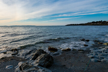 Rocks in the foreground with ocean waves rolling over them. The blue ocean has small ripples over the water.  The sky is blue with white wispy clouds. The land at the horizon is a light orange color. 