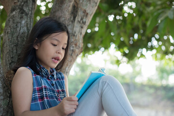  little asian girl reading a book under big tree. children and science. blurred background. learning the imagination and dreams of rural child. studying at home.