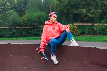 Young girl with short multicolored hair sits with skateboard in skatepark