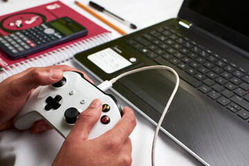 Young man plays a video game with a wired control on his laptop while leaving school aside in times...