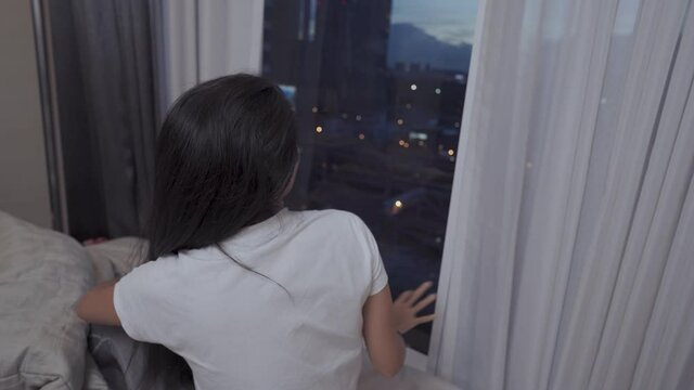 Young Asian lady Sit down tie hair after wake up on the bed, dust sundown evening time, afternoon nap getting up next to bedroom window and curtain, high rise condominium with city urban highway view