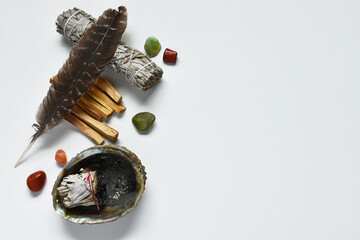 A top view image of white sage, palo santo, healing crystals, and a sacred feather on a white...