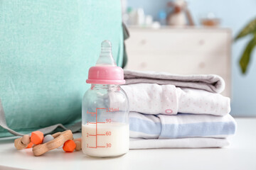 Bottle of milk for baby with clothes and bag on table in room
