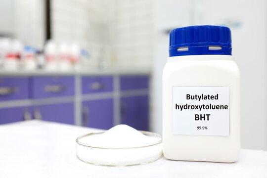 Selective focus of a bottle of BHT or butylated hydroxytoluene chemical antioxidant beside a petri dish with white crystalline powder substance. White laboratory background with copy space.