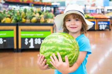 Fototapeta premium Kid holding watermelon in supermarket against food stall. Smiling positive boy grocery shopping at the supermarket.
