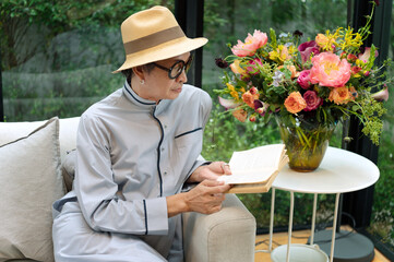 Senior LGBT reads a book in sofa next to folower vase containing colorful flower (rose, lily and peony) with clam and relaxing gesture in family living room