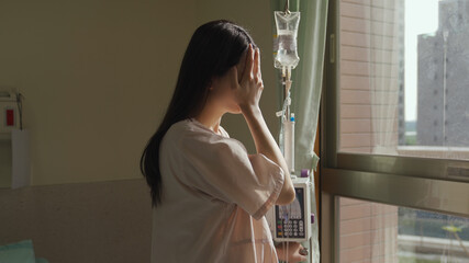crying woman standing with drip stand in front of window is thinking about her illness and wiping...