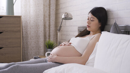 portrait of asian korean mother-to-be resting in bed feeling her baby moving in big naked abdomen...