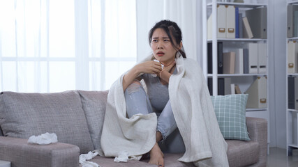 coughing chinese infected girl in self-quarantine at home and covered with a blanket huddling up on couch resting.