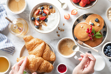 breakfast with croissants, granola with yogurt, pancakes, fresh berries, honey, nuts and coffee on a white marble background