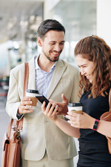 Laughing young coworkers drinking take away coffee and watching funny videos on smartpphone during break