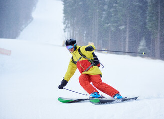 Fototapeta na wymiar Front view of young male skier downhill skiing and doing carve turn on high snowy slope. Skiing during snowfall in mountains. Single descent. Winter foggy background. Active sporty lifestyle concept.