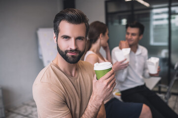 Bearded male enjoying coffee, his male and female colleagues sitting on table, having lunch
