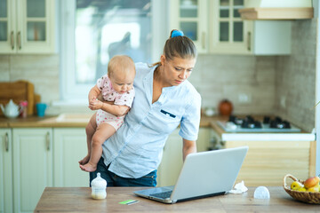 A woman housewife at home in the kitchen sits at the table with a laptop and a little baby. Communicates, relaxes, works from home and shop online