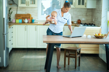 A woman housewife at home in the kitchen sits at the table with a laptop and a little baby. Communicates, relaxes, works from home and shop online
