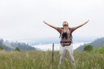 Young man with backpack standing on top mountain with raised hands at morning sunrise with foggy mountain landscape