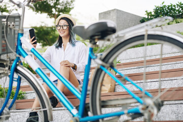 Fototapeta na wymiar Happy young Asian woman sitting on steps and taking selfie after bicycle ride