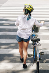 Young woman in white shirt and denim shorts walking next to bicycle when crossing road
