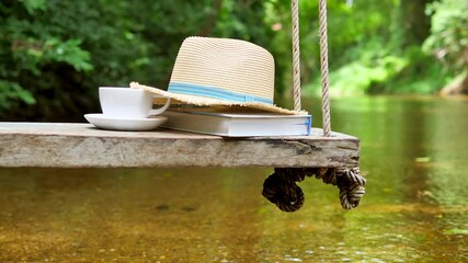Old wood swing hanged on a tree and A river flows in beautiful scene with book, Cup of coffee and hat. ( Water stream )