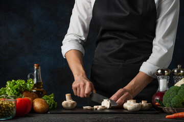 Chef in white shirt and black apron cuts mushrooms on a dark table isolated on black dark background. Different vegetables are on the table. Tasty dish. Restaurant meals. Food concept.Hands with knife