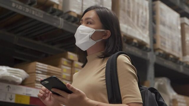 Asian woman wearing mask walking in warehouse and use smart phone for check price.