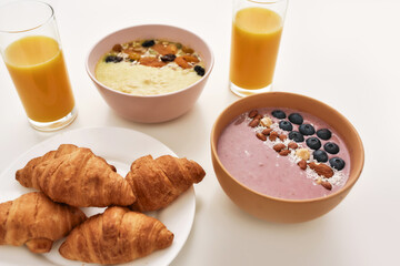 Close up of smoothie bowl with fresh berries, orange juice in glass and croissants on a plate isolated over white background
