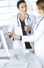 Two female physicians are discussing medical therapy, while standing at the table in a clinic office. Doctors use pc computer at work. Teamwork in medicine