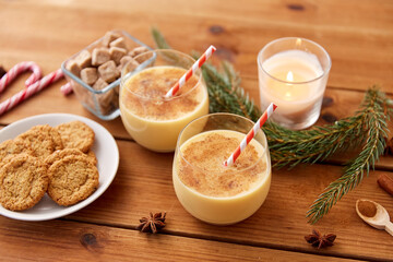 christmas and seasonal drinks concept - glasses of eggnog with oatmeal cookies, candy canes, sugar, fir tree branches and candle burning on wooden background