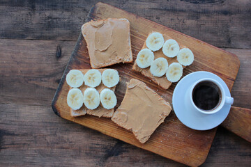 Cup of coffee and peanut butter banana toasts on wooden background. Slices of whole wheat bran bread with peanut paste on a cutting board. Healthy breakfast