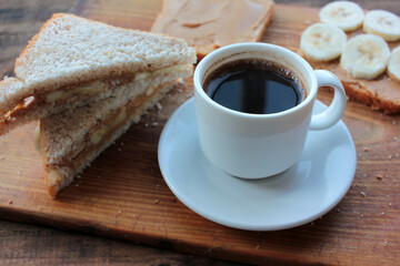 Obraz na płótnie Canvas Cup of coffee and peanut butter banana toasts on wooden background. Slices of whole wheat bran bread with peanut paste on a cutting board. Healthy breakfast