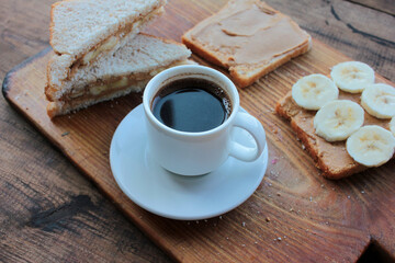 Cup of coffee and peanut butter banana toasts on wooden background. Slices of whole wheat bran bread with peanut paste on a cutting board. Healthy breakfast