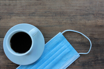 Cup of coffee and disposable medical face mask on wooden table from above. New normal. 