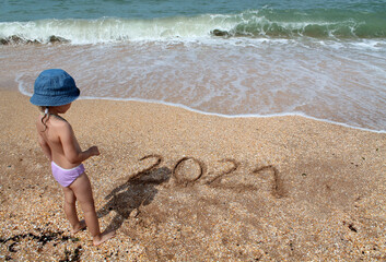 A little girl in a blue Panama hat stands near the inscription on the sand 