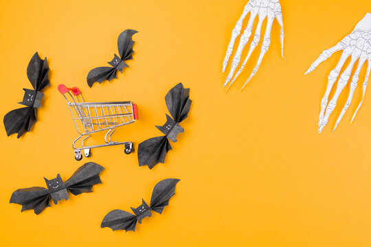 The customer's toy cart is located in the middle of paper decorative bats, to which the skeleton's hands reach. Orange background. Copy space. Flat lay. The concept of Halloween and holiday shopping