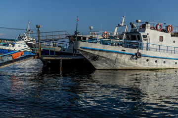 white vessel ship boat with blue stripes on pier in bay of lake baikal in light of sun with reflections in water
